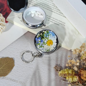 Peut inclure: Un pilulier rond argenté avec un motif floral sur le couvercle. Le pilulier est ouvert, révélant des pilules blanches. Le couvercle présente un arrangement de fleurs pressées blanches, jaunes et bleues. Un porte-clés argenté est attaché.