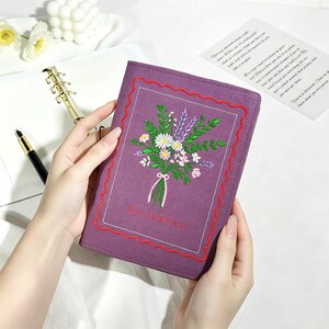 May include: A purple fabric-covered notebook featuring an embroidered bouquet of white, pink, and purple flowers with green leaves. The cover is framed by a red scalloped stitched border and includes small embroidered text at the bottom.