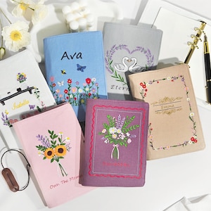 May include: A collection of six fabric-covered notebooks featuring embroidered designs including floral bouquets, a swan heart, and personalized names like Ava, Isabella, and Emma. The covers come in various colors such as light blue, pink, purple, and beige.