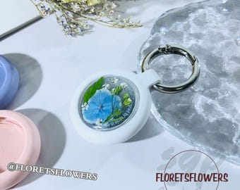 May include: White keychain with a clear round charm containing pressed blue and white flowers. The charm has the text "Made in Hawaii by Florets Flowers" printed on it.