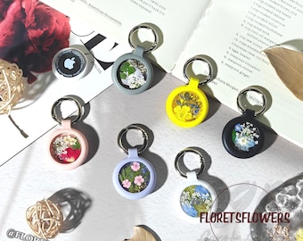 May include: A collection of keychains in various colors, each featuring a clear circular window filled with dried flowers. The keychains have silver-colored rings and are arranged on a light-colored surface. The brand name "FLORETSFLOWERS" is visible.