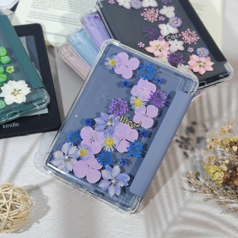 K&ouml;nnte beinhalten: Transparente Kindle-H&uuml;llen mit gepressten lila, blauen und gelben Blumen. Die H&uuml;lle hat eine blaue Abdeckung und das Wort "Alice" in Goldbuchstaben. Weitere H&uuml;llen mit floralen Designs sind sichtbar.