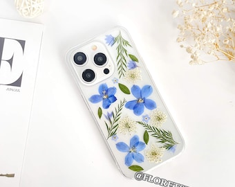 May include: A clear phone case with a design of pressed blue and white flowers. The case has a white background and the flowers are arranged in a random pattern. The case is for an iPhone with a triple lens camera. The text "@floretsflowers" is printed on the bottom of the case.