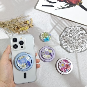 May include: A clear smartphone case with a blue circular phone grip featuring pressed blue and white flowers. Three additional circular phone grips are displayed nearby, showcasing various pressed floral designs in blue, purple, and pink color palettes.