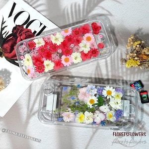 Op de afbeelding: Twee doorzichtige Switch-gameconsole-hoesjes gevuld met kleurrijke gedroogde bloemen. Eén hoesje heeft rode, roze en witte bloemen; de andere heeft blauwe, gele en witte bloemen. Op een witte ondergrond, met een Vogue-tijdschrift.