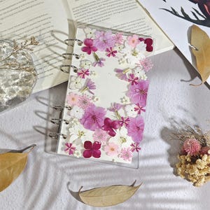 Può includere: Un raccoglitore rettangolare in acrilico trasparente con una collezione di fiori pressati nelle tonalità del rosa, viola e bianco disposti sulla copertina. Il raccoglitore presenta tre anelli metallici argentati sul lato sinistro.