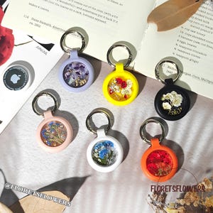 May include: A collection of colorful AirTag holders with dried flowers encased in clear resin. The holders come in various colors, including lavender, yellow, black, pink, white, and orange, each with a silver keyring. The text "FLORETSFLOWERS" is visible.