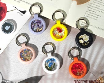 May include: A collection of colorful AirTag holders with dried flowers encased in clear resin. The holders come in various colors, including lavender, yellow, black, pink, white, and orange, each with a silver keyring. The text "FLORETSFLOWERS" is visible.