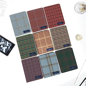 Könnte beinhalten: Eine Sammlung von neun rechteckigen Stoffhüllen mit verschiedenen Karomustern und Tartans in Farben wie Blau, Rot, Grün, Braun und Violett. Jede Hülle hat ein kleines dunkelblaues rechteckiges Etikett mit der Aufschrift Holland & Sherry.