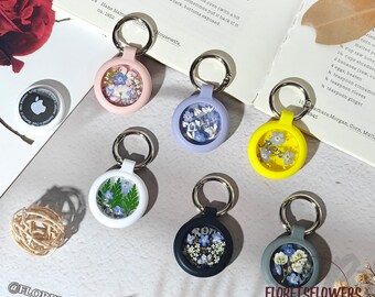 May include: A collection of AirTag holders in various colors, each featuring dried flowers encased within a clear, circular frame. The holders have silver key rings. Some holders have text that reads "Rex" and "RO". The background includes a book and flowers.