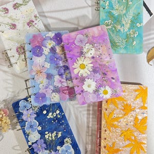 Puede incluir: Una colección de cinco cuadernos rectangulares de resina con flores y hojas prensadas incrustadas. Cada cuaderno tiene una encuadernación de anillas metálicas en el lateral. Las portadas muestran motivos florales en tonos azules, morados y dorados.