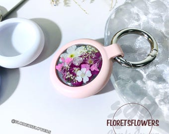 May include: A pink keychain with a clear round charm containing pressed flowers. The charm is attached to a silver metal ring and a pink strap. The text "FloretsFlowers" is visible in the image.