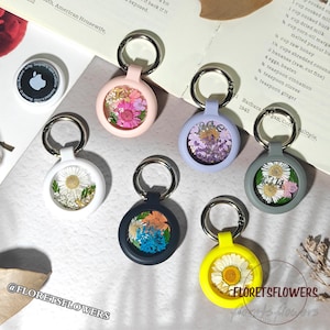 May include: A collection of keychains with dried flowers encased in clear resin. The keychains come in various colors, including pink, white, blue, gray, and yellow. Each keychain has a silver ring. The text "FLORETSFLOWERS" is visible.