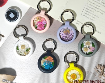 May include: A collection of keychains with dried flowers encased in clear resin. The keychains come in various colors, including pink, white, blue, gray, and yellow. Each keychain has a silver ring. The text "FLORETSFLOWERS" is visible.