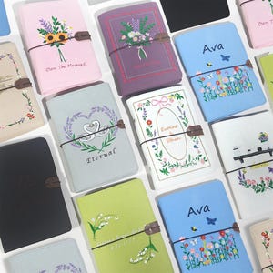 May include: A collection of rectangular fabric-covered journals featuring various embroidered designs including floral bouquets, swans, and butterflies, with some covers personalized with names like Ava, Evonne, and Ethan, and phrases like Own The Moment.