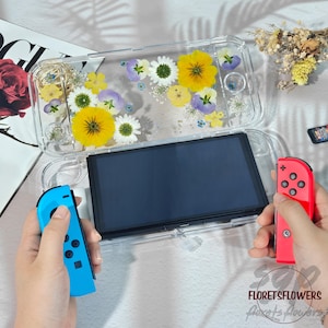May include: Clear protective case for a handheld gaming device, adorned with pressed yellow, white, and purple flowers. The device has a black screen and is held with a blue and red controller.