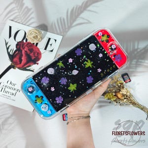 May include: A clear Switch case with a black background, decorated with pressed purple and green flowers, and silver glitter. The case has red and blue accents. The name "Bruce" is written on the case.