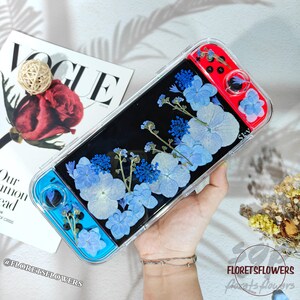 May include: A clear gaming device case with blue and red controllers, decorated with pressed blue flowers against a black backdrop. The case is held in a hand, with a magazine and a rose visible in the background.