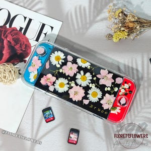 May include: A clear protective case for a gaming console, featuring a black interior adorned with pressed pink and white flowers. The case has blue and red accents and the name "Bruce" is visible.