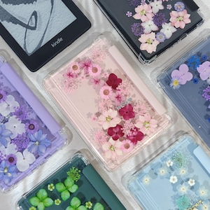 K&ouml;nnte beinhalten: Eine Sammlung klarer H&uuml;llen f&uuml;r elektronische Ger&auml;te, die jeweils mit gepressten Blumen in verschiedenen Farben verziert sind. Die H&uuml;llen zeigen eine Vielzahl von Blumenarrangements, darunter rosa, lila und wei&szlig;e Blumen, die einen dekorativen und k&uuml;nstlerischen Look erzeugen.