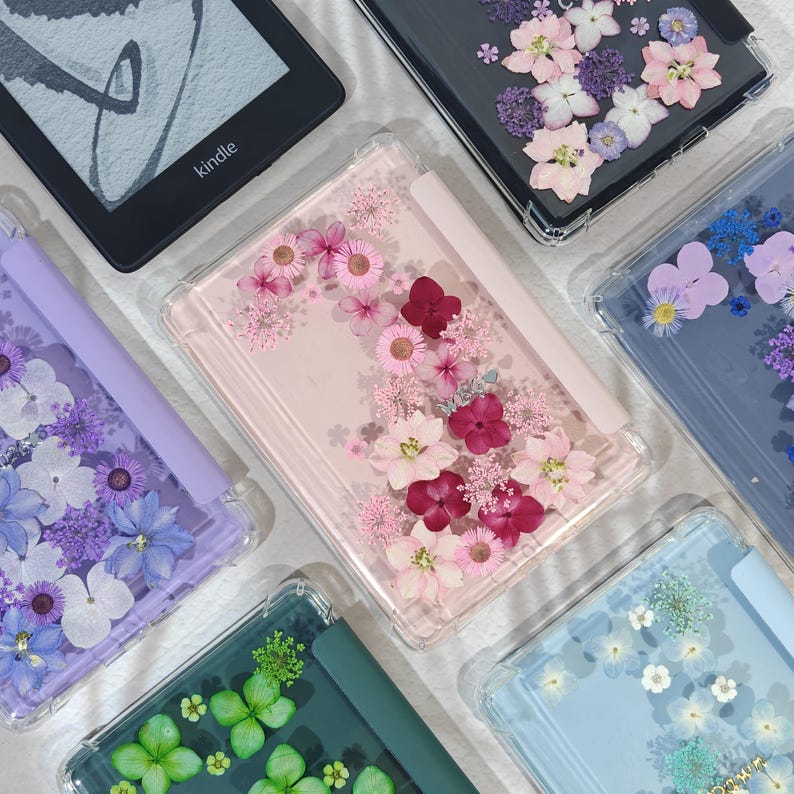 K&ouml;nnte beinhalten: Eine Sammlung klarer H&uuml;llen f&uuml;r elektronische Ger&auml;te, die jeweils mit gepressten Blumen in verschiedenen Farben verziert sind. Die H&uuml;llen zeigen eine Vielzahl von Blumenarrangements, darunter rosa, lila und wei&szlig;e Blumen, die einen dekorativen und k&uuml;nstlerischen Look erzeugen.