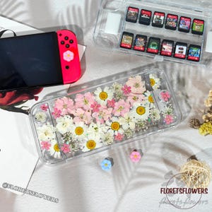 May include: A clear case for a Nintendo Switch Lite, filled with pink and white pressed flowers and glitter. A pink and red Switch controller is visible, along with a clear case holding game cartridges. The case is decorated with the text "FLORETSFLOWERS".