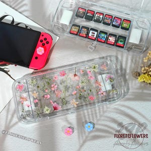 May include: Clear plastic Nintendo Switch case with embedded pink and white flowers. A second clear case holds game cartridges. A pink and black Switch console is visible. The text "FLORETSFLOWERS" is in the bottom right corner.