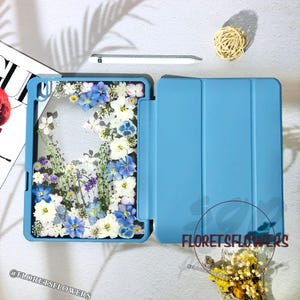 Puede incluir: Una funda azul claro para iPad con una ventana transparente llena de flores prensadas azules, blancas y moradas. La funda está abierta, revelando la pantalla. El texto "FLORETSFLOWERS" es visible en la funda.
