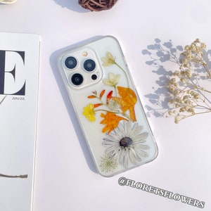 May include: A white iPhone case with a pressed flower design. The flowers are white, yellow, orange, and red. The case has a clear back and a white border. The text "@floretsflowers" is printed on the bottom of the case.