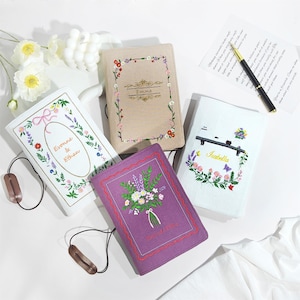 May include: Four rectangular fabric book covers featuring embroidered floral designs and personalised names. One cover is beige with a floral border, one is white with a floral oval, one is white with a floral wreath, and one is purple with a bouquet.