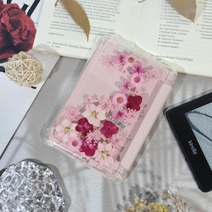 May include: A clear phone case with a pink border, filled with an arrangement of pink and burgundy dried flowers. The case is open, revealing a light pink interior. A Kindle e-reader is visible in the background.