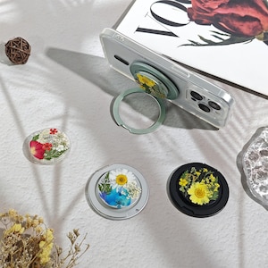 May include: Three circular phone ring holders featuring pressed flower designs. One holder is attached to a clear phone case, while two others are separate. The designs include red, white, and yellow flowers encased in clear, silver, and black frames.