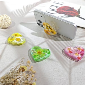May include: Four heart-shaped phone grips featuring embedded dried flowers. One grip is attached to a white smartphone, while three others are displayed separately. The designs include yellow daisies, green foliage, and pink floral arrangements.