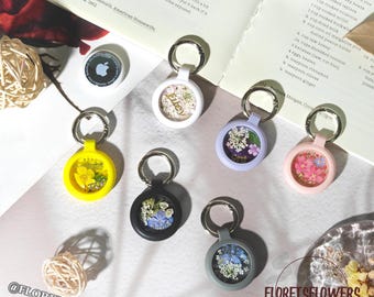 May include: A collection of keychains with dried flowers encased in clear, round holders. The keychains come in various colors, including yellow, black, gray, pink, and purple. Each keychain has a silver ring. The text "FLORETSFLOWERS" is visible.