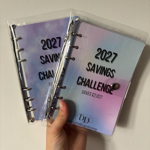 May include: Two clear plastic binders with metal rings, each containing a pastel purple and blue gradient insert. The front cover features the text 2027 SAVINGS CHALLENGE, SAVES £2,027, and a logo with the text DD delightdesignsbymeg at the bottom.