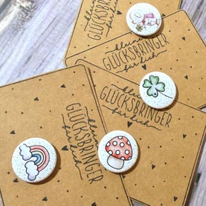 Könnte beinhalten: Eine Sammlung kleiner, runder, handbemalter Anstecknadeln mit verschiedenen Designs, darunter ein Regenbogen, ein Pilz, ein Kleeblatt und ein Hase. Die Anstecknadeln liegen auf braunen Karten mit dem Text "kleine GLÜCKSBRINGER für dich".