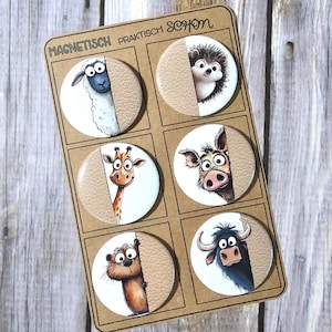Könnte beinhalten: Ein Set aus sechs runden Magneten mit Cartoon-Tierillustrationen. Jeder Magnet zeigt einen Teil eines Tieres, darunter ein Schaf, ein Igel, eine Giraffe, eine Kuh, ein Otter und ein Bulle. Die Magnete befinden sich auf einer braunen Kartonrückseite mit deutschem Text.