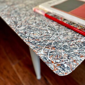May include: A white table with a black, gray, orange, and white abstract paint design. Two books are stacked on the table, with a red pencil on top of the books. The top book has the text "TASCHEN" on the cover.
