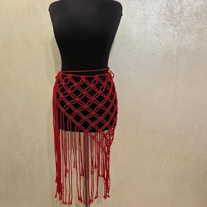 May include: A red macrame skirt with a matching choker displayed on a black mannequin. The skirt features a geometric pattern and long fringe. The choker is a simple band. The mannequin is on a silver stand.