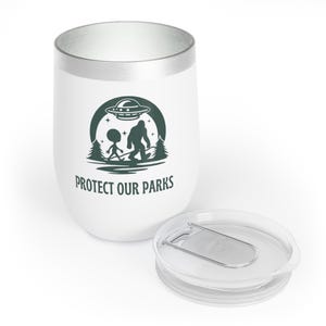 May include: White stainless steel tumbler with a green graphic of a Bigfoot and an alien walking in a forest with a UFO above them. The tumbler has a silver lid and the text "PROTECT OUR PARKS" is printed on the side.