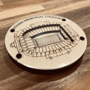 May include: A wooden coaster featuring a black and white outline of Kenan Memorial Stadium, home of the University of North Carolina Tar Heels football team. The coaster has a black border and the words "Kenan Memorial Stadium" are printed around the top edge.