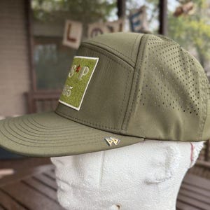 May include: Olive green baseball cap with a flat brim and perforated side panels. The cap features a rectangular patch with the letters "S P" and the number "95". A small silver emblem is on the side.