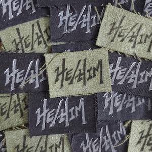 May include: Close-up of a pile of fabric patches with the text "He/Him" printed in silver on a black and green background.