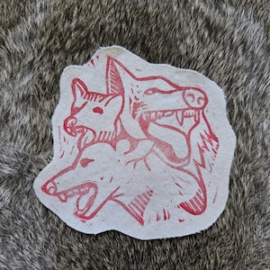 May include: A light-colored fabric patch featuring a red ink block print illustration of three snarling wolf heads arranged in a cluster. The design displays sharp teeth, pointed ears, and textured linework details on a plain, off-white textile base.