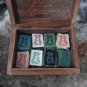 May include: A wooden box with a hinged lid, open to reveal nine small fabric squares. Each square features an embroidered design of a seated figure with a crescent moon above its head. The squares are in various colors, including teal, green, black, white, and red.
