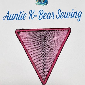 May include: A triangular embroidered patch featuring a light pink thread pattern with a dark pink border. Above the patch, the text "Auntie K-Bear Sewing" is printed in blue, accompanied by a small graphic of a bear using a sewing machine.