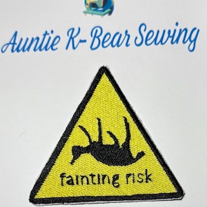 May include: A triangular embroidered patch with a yellow background and a thick black border. The patch features a black silhouette of a person lying on their back with limbs raised, and the black text "fainting risk" printed below.