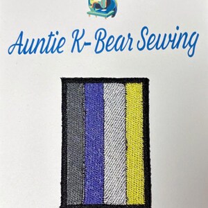 Könnte beinhalten: Ein rechteckiger gestickter Aufnäher mit vier vertikalen Streifen in Grau, Lila, Weiß und Gelb, umrandet von einem schwarzen Rand. Über dem Aufnäher steht der Text "Auntie K-Bear Sewing" in blauer Schrift, ergänzt durch ein kleines Bärenlogo.