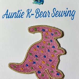 Peut inclure: Écusson de dinosaure rose brodé avec des pois colorés et une bordure dorée. L'écusson présente le texte "Auntie K-Bear Sewing" et un ours de dessin animé avec une machine à coudre.