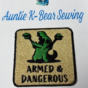 Op de afbeelding: Geborduurde patch met de tekst "ARMED & DANGEROUS" in zwart op een gouden achtergrond. De patch toont een groen silhouet van een wezen dat twee objecten vasthoudt. De patch is vierkant met een zwarte rand. De tekst "Auntie K-Bear Sewing" staat boven de patch.